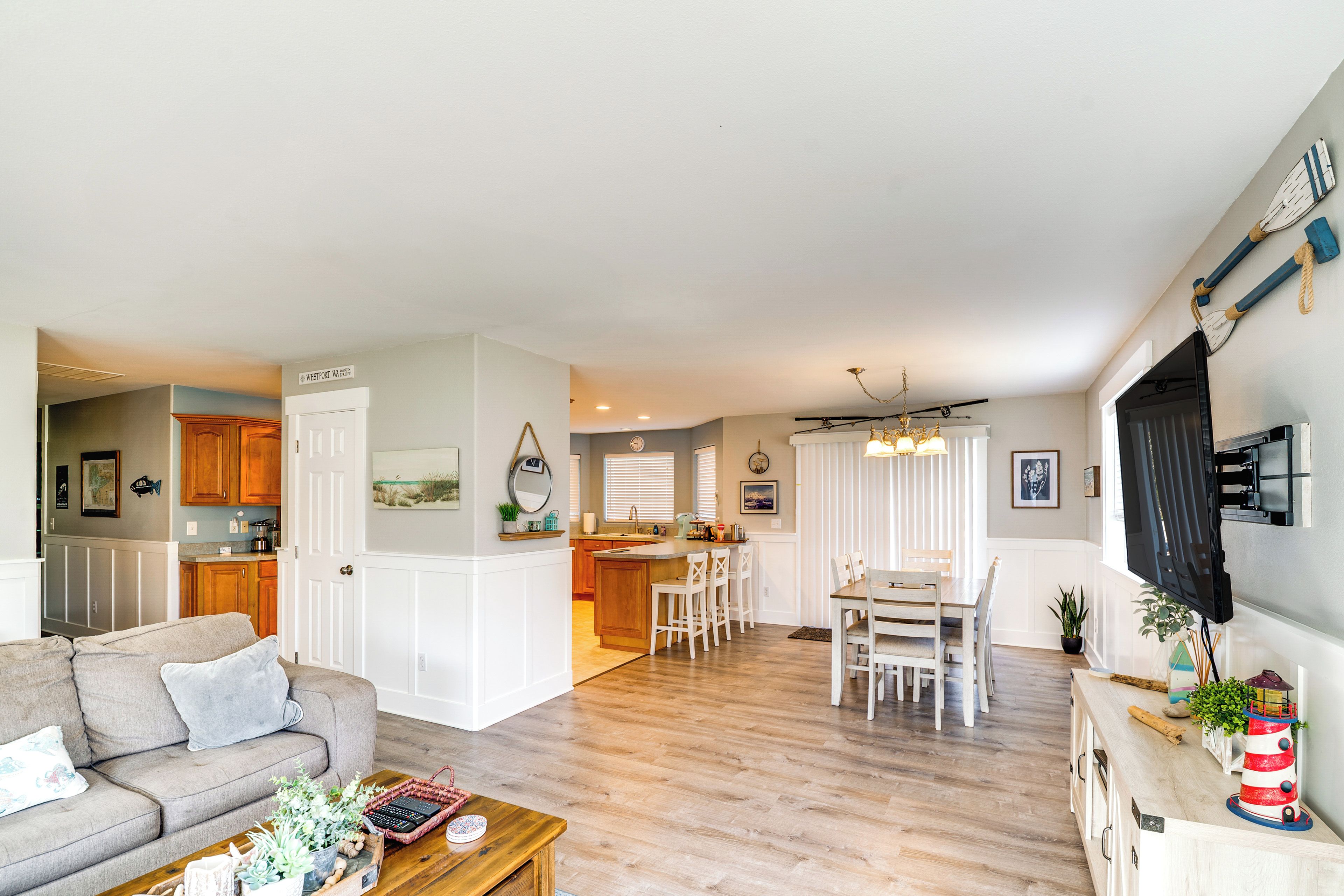A spacious living area with a sofa, coffee table, and a dining table with chairs. A kitchen with wooden cabinets and a dining area with a chandelier are visible.