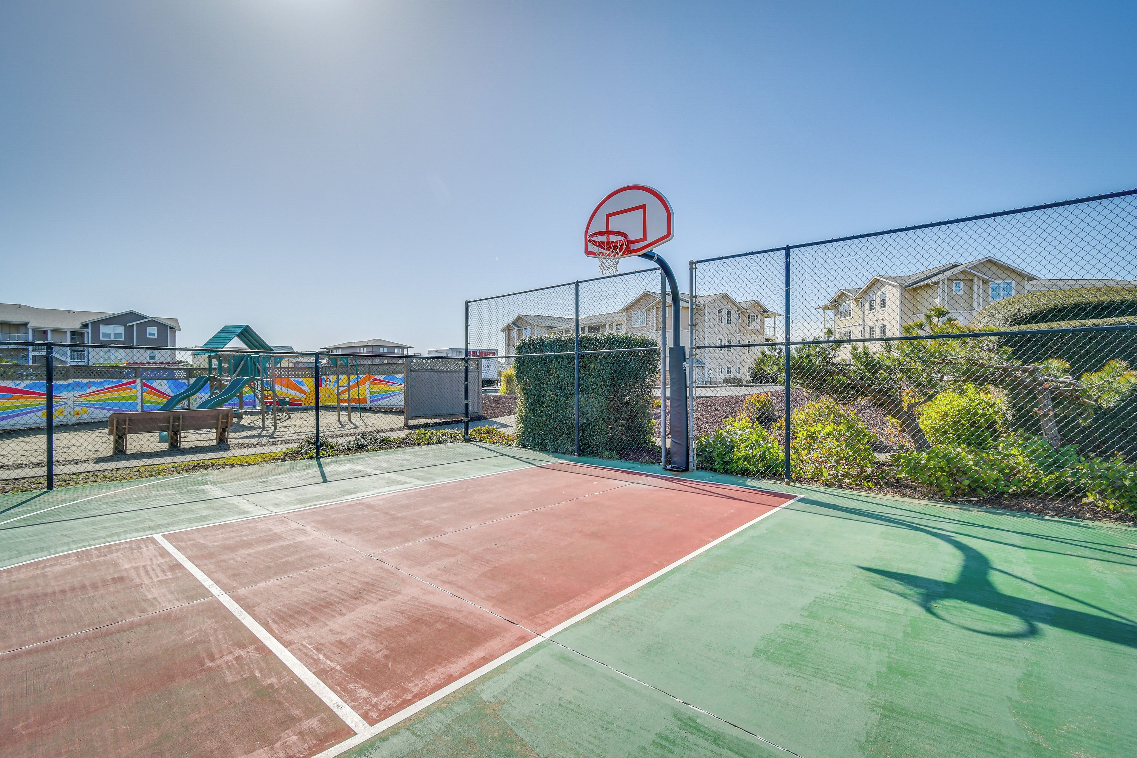 An outdoor basketball court with a fence, a bench, and a colorful playground structure in the background.