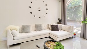 A modern living room with a white sectional sofa, a large wall clock, a coffee table with pebbles, and a large window with curtains. - Modern & Pet-friendly Austin Vacation Rental! (Austin)