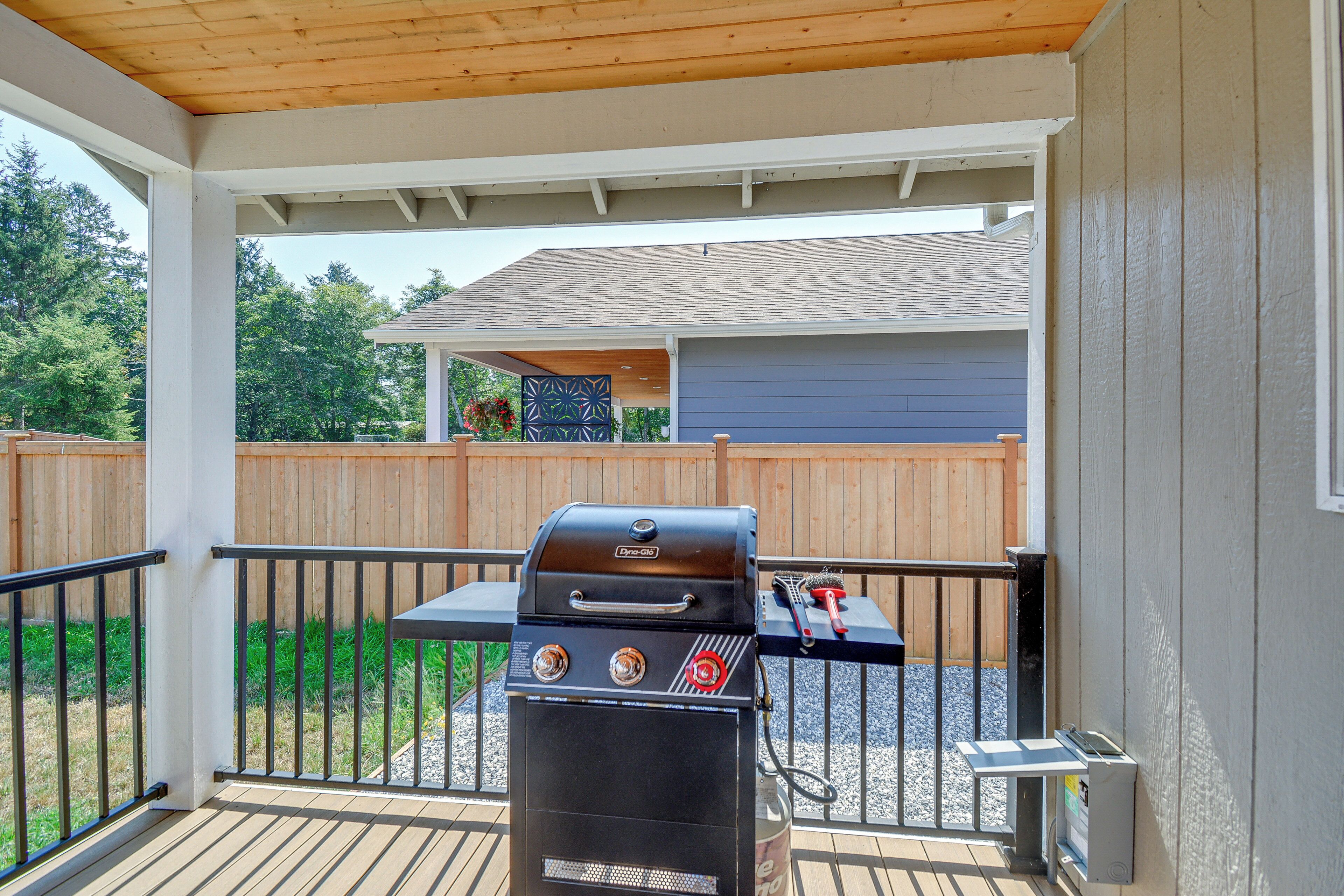 Een overdekt terras buiten met een barbecue, een tafel met borstel en een houten omheining op de achtergrond.