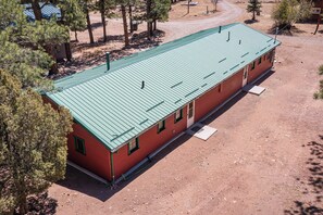A long, single-story building with a green roof, surrounded by trees and a dirt ground. - Secluded Stonewall Cabin w/ Mountain View! (Weston)