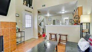 A living room with a sofa, a coffee table, a TV mounted on the wall, and a kitchen area with a counter and stools.