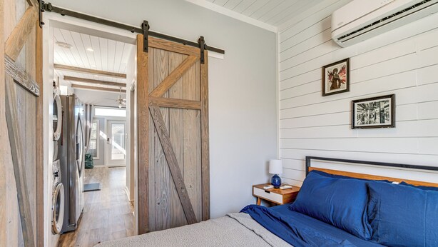 A bedroom with a bed, a nightstand, a wall-mounted air conditioner, and a sliding barn door leading to a laundry area. - Austin Tiny Home: Fireplace, Shared Pool & Hot Tub (Austin)