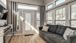 A modern living room with a grey sofa, a flat-screen TV, and large windows with blinds. - Austin Tiny Home: Fireplace, Shared Pool & Hot Tub (Austin)