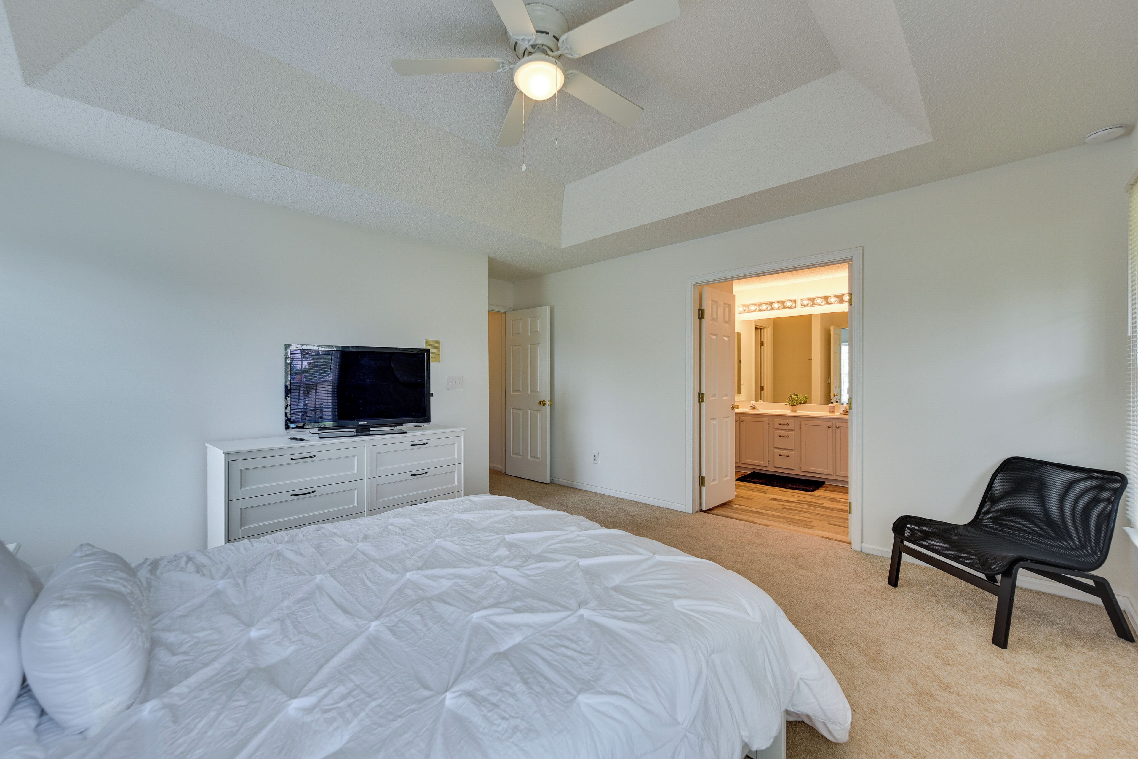 A bedroom with a bed, a TV on a dresser, a ceiling fan, and a bathroom visible through an open door.
