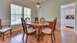Een eethoek met een houten tafel en stoelen, een hanglamp en uitzicht op een keuken door een open deur.