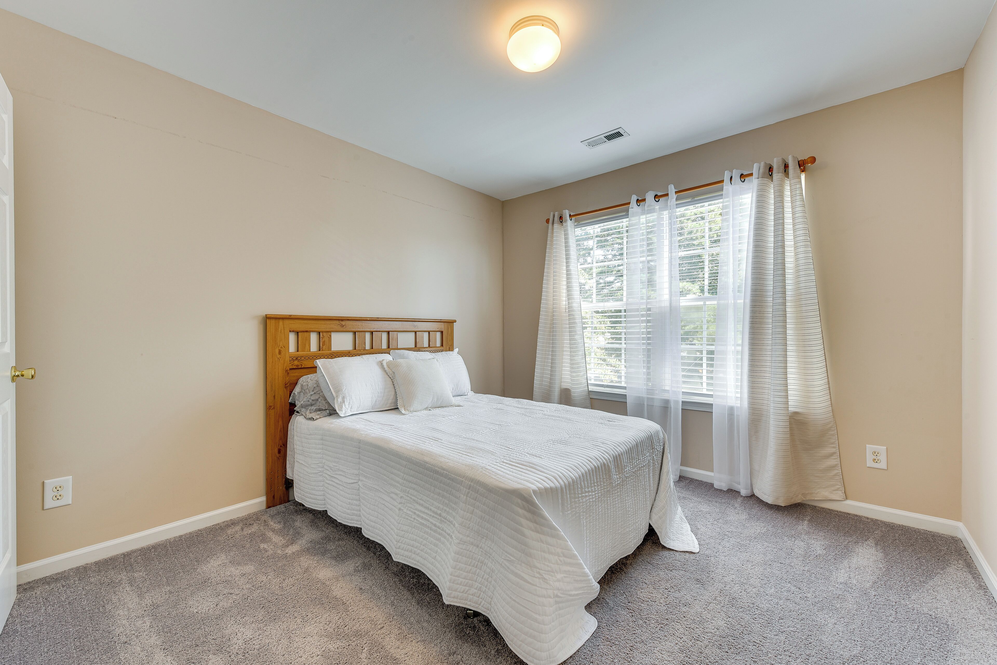 Un dormitorio con cama de madera, paredes color beige y una ventana con cortinas blancas.