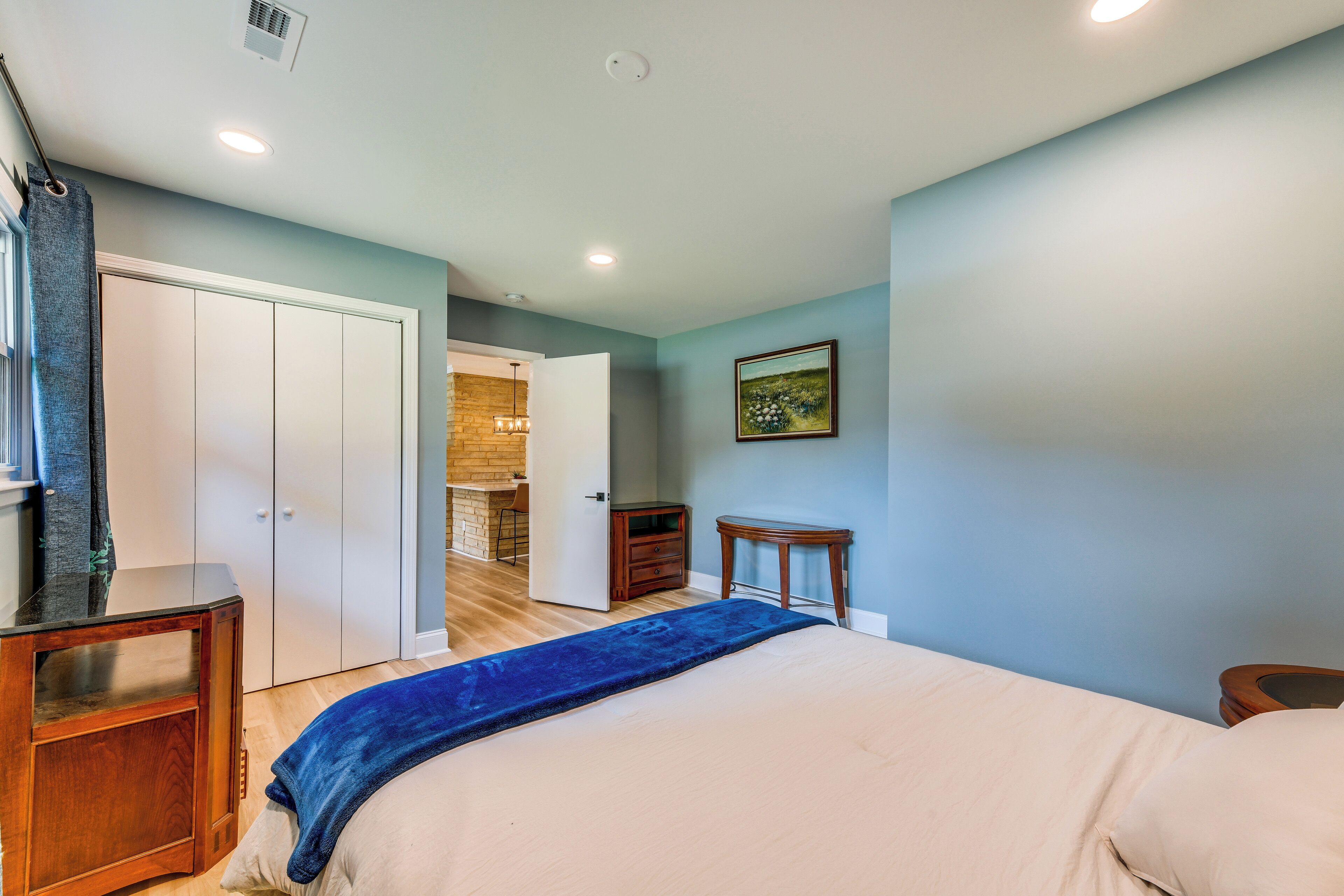 Un dormitorio con una cama, una mesita de noche de madera, un aparador y una puerta que da a una zona de cocina.