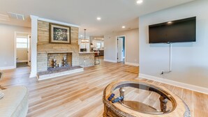 A spacious living room with hardwood floors, a fireplace, a mounted TV, and a glass coffee table. - Charming Charlotte Home: 2 Mi to Downtown! (Charlotte)