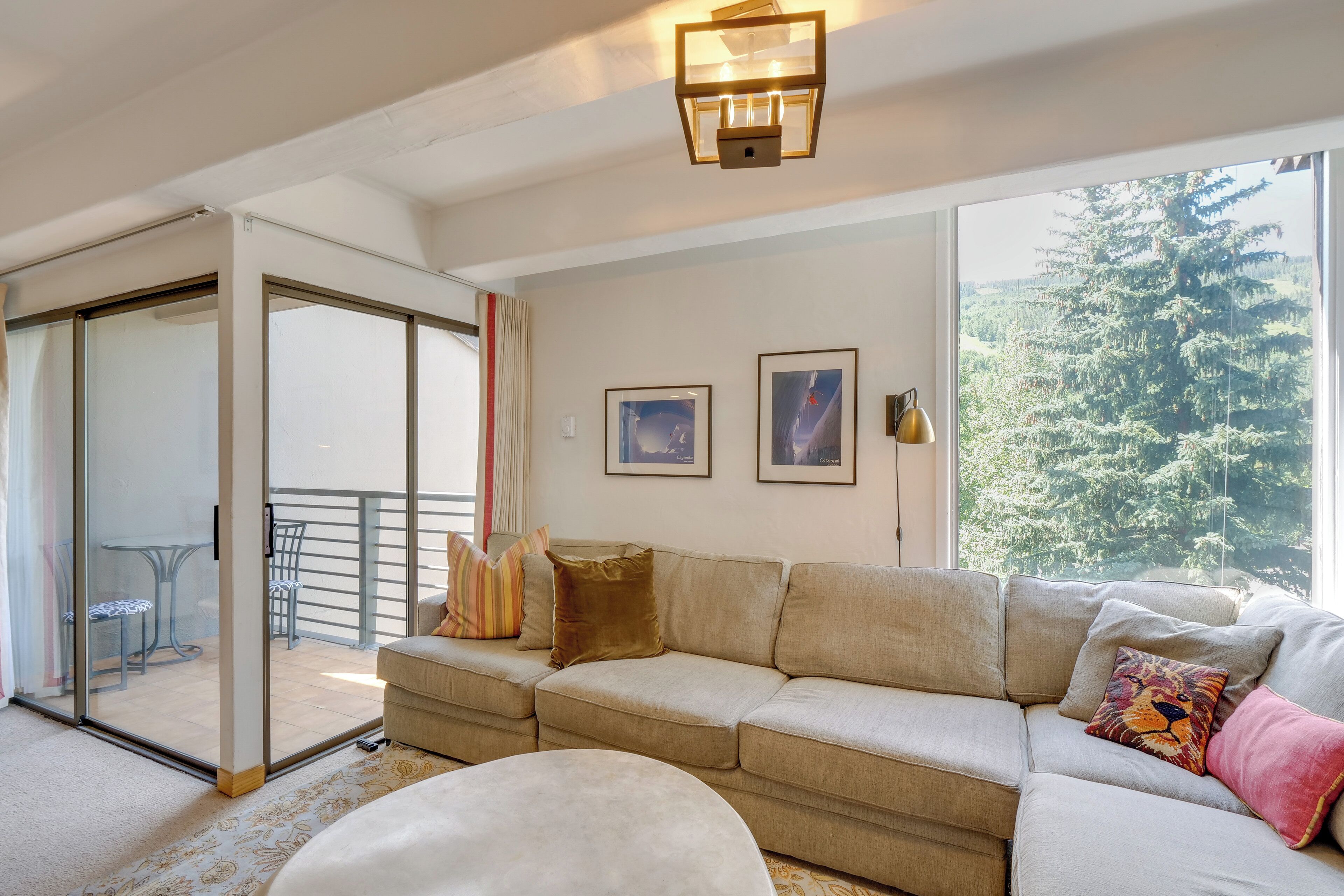 A living room with a beige sofa, a round ottoman, and a glass door leading to a balcony.