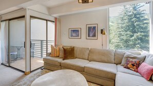 A living room with a beige sofa, a round ottoman, and a glass door leading to a balcony. - Vail Top-floor Condo w/ Deck & Mountain Views! (Vail)