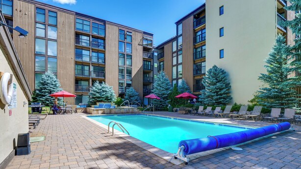 A swimming pool area with lounge chairs and umbrellas, surrounded by multi-story buildings with balconies. - Vail Top-floor Condo w/ Deck & Mountain Views! (Vail)