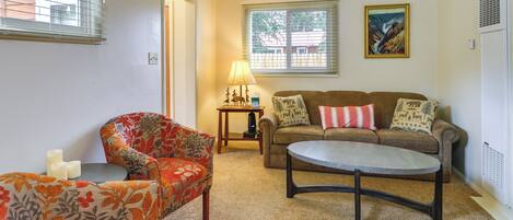A living room with a sofa, armchair, and coffee table. There is a lamp on a side table, and a painting on the wall.
