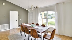 Un coin repas comprenant une table en bois, six chaises, un lustre et une grande fenêtre agrémentée de rideaux.