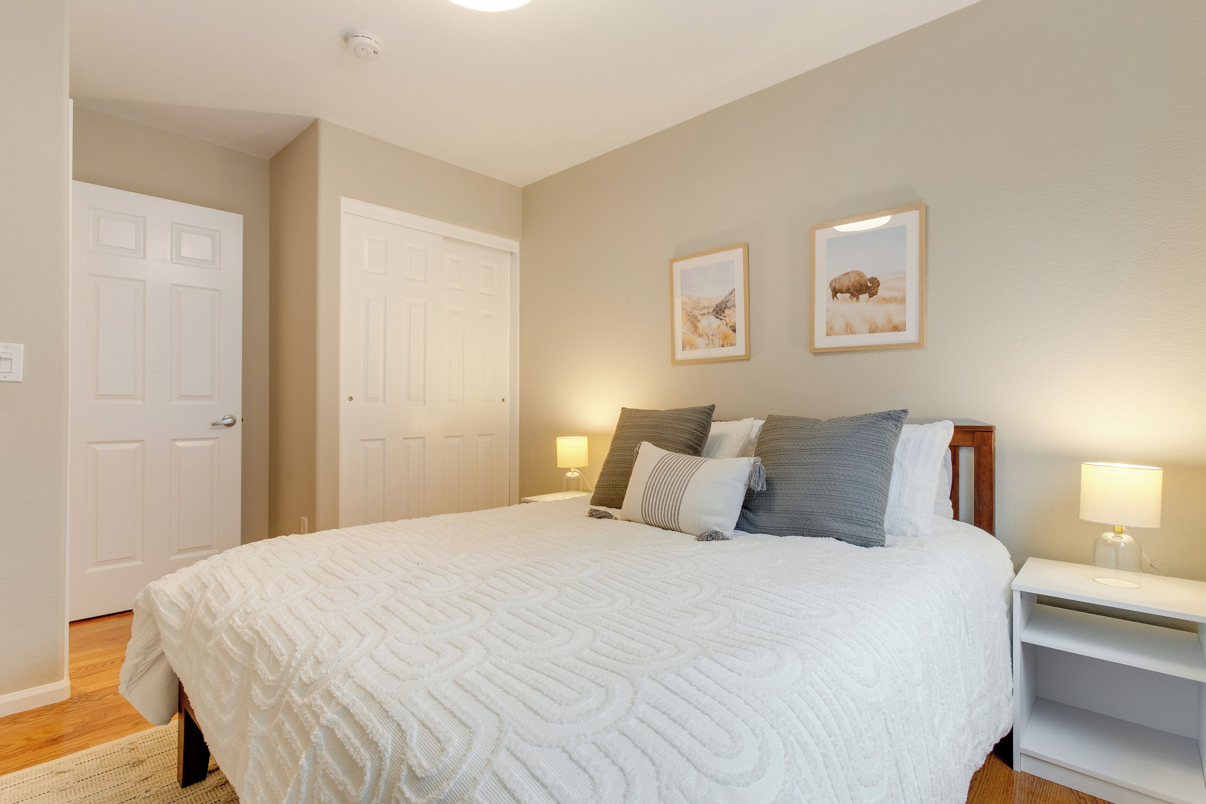 A bedroom with a large bed, two bedside tables with lamps, a door, and two framed pictures on the wall.