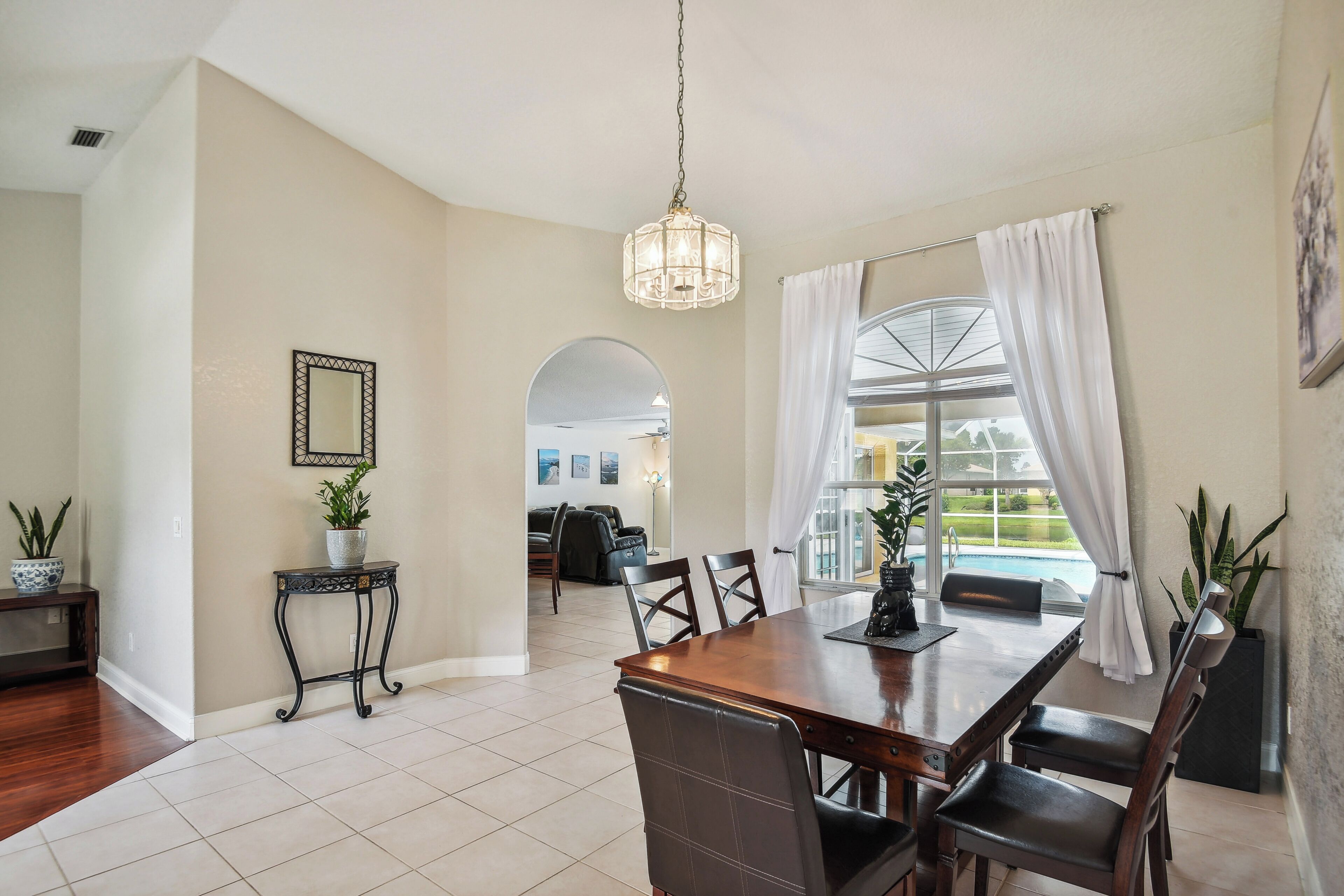 A dining area with a wooden table and chairs, a hanging light fixture, and a view of a pool through a window with white curtains.