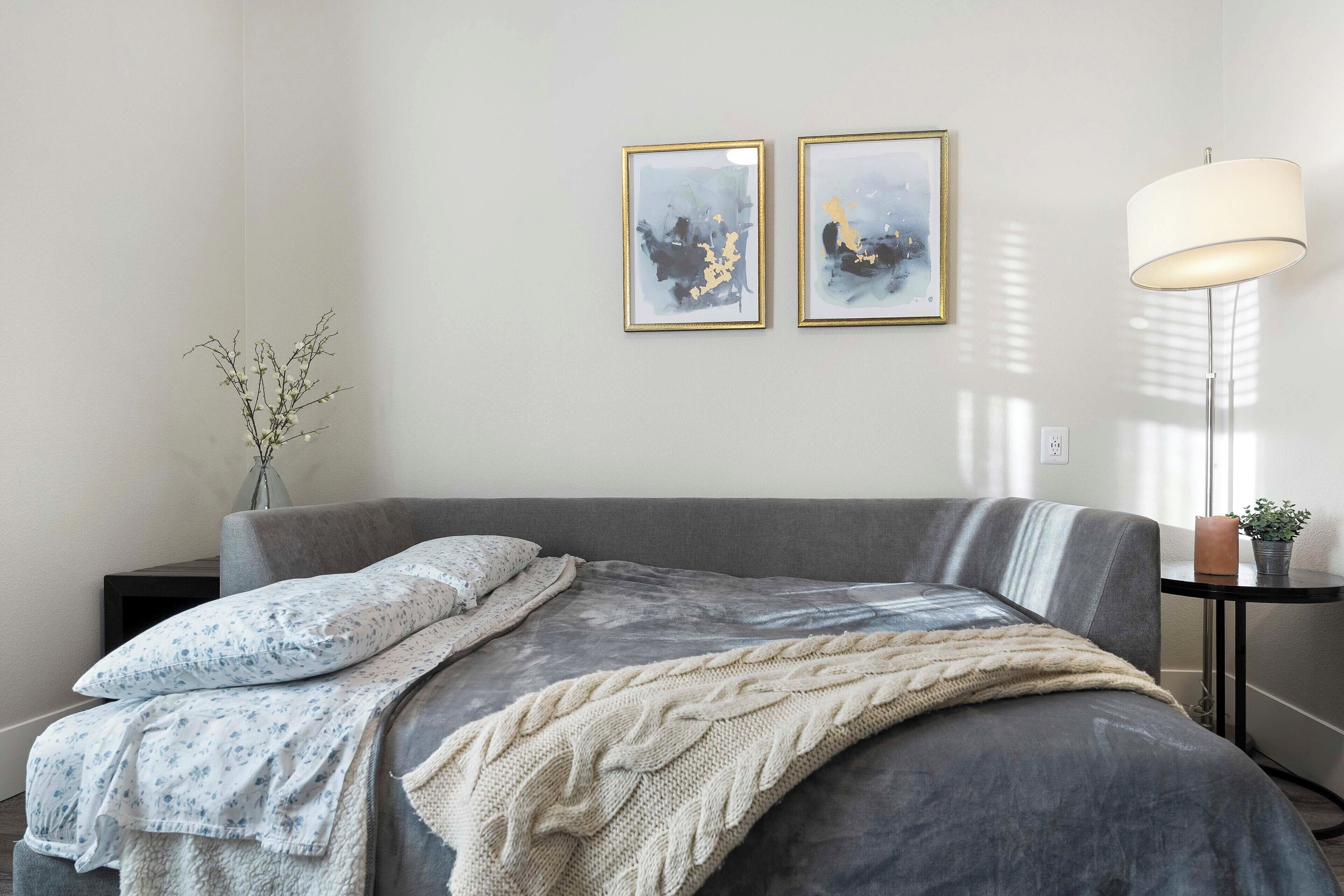 Ein modernes Schlafzimmer mit einer grauen Schlafcouch, einer weiß-blauen Blumendecke, einer beigen Strickdecke und zwei abstrakten Bildern an der Wand.