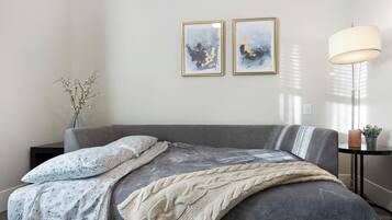 Ein modernes Schlafzimmer mit einer grauen Schlafcouch, einer weiß-blauen Blumendecke, einer beigen Strickdecke und zwei abstrakten Bildern an der Wand.
