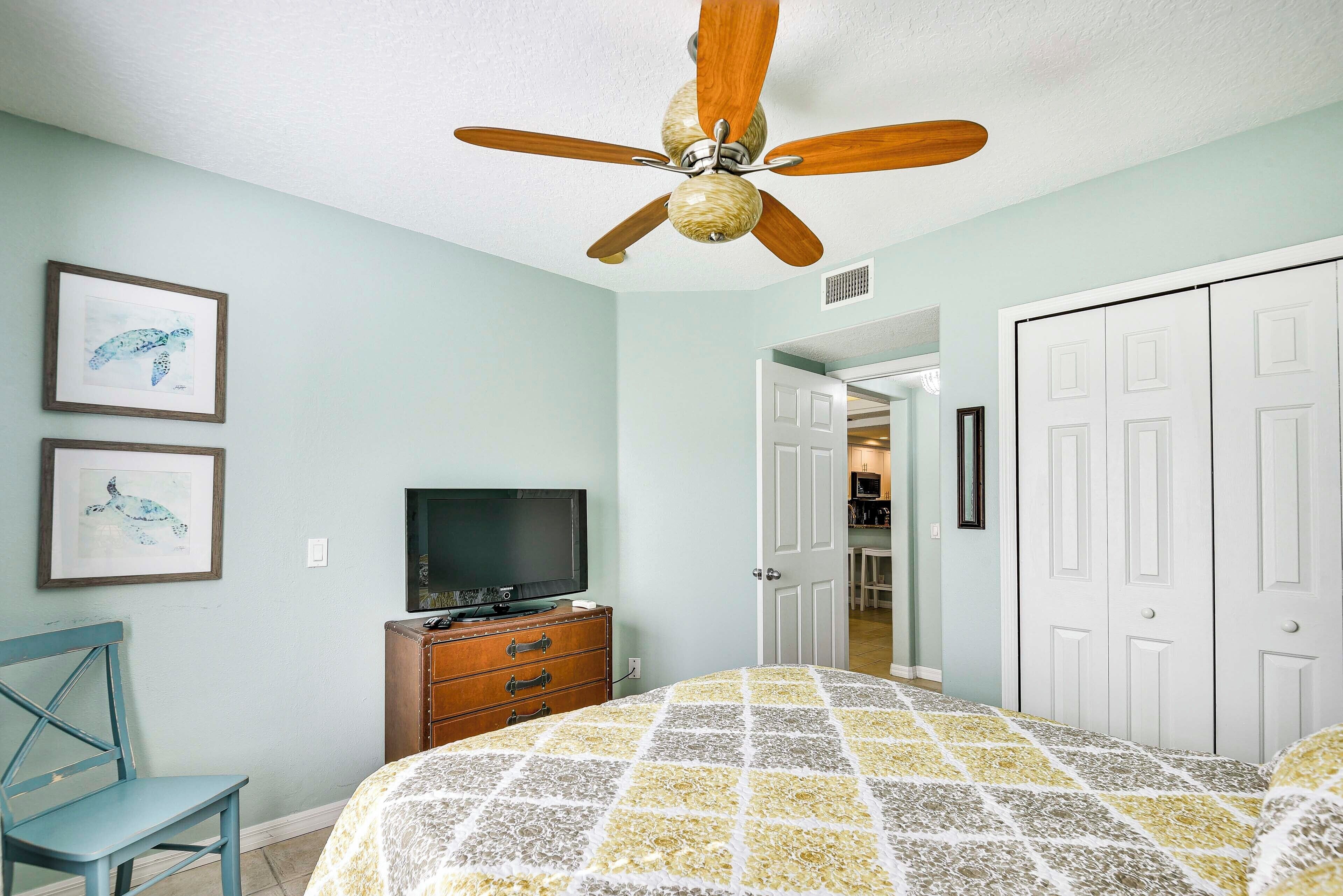 A bedroom with a bed, a ceiling fan, a dresser, a television, and framed artwork on the wall.