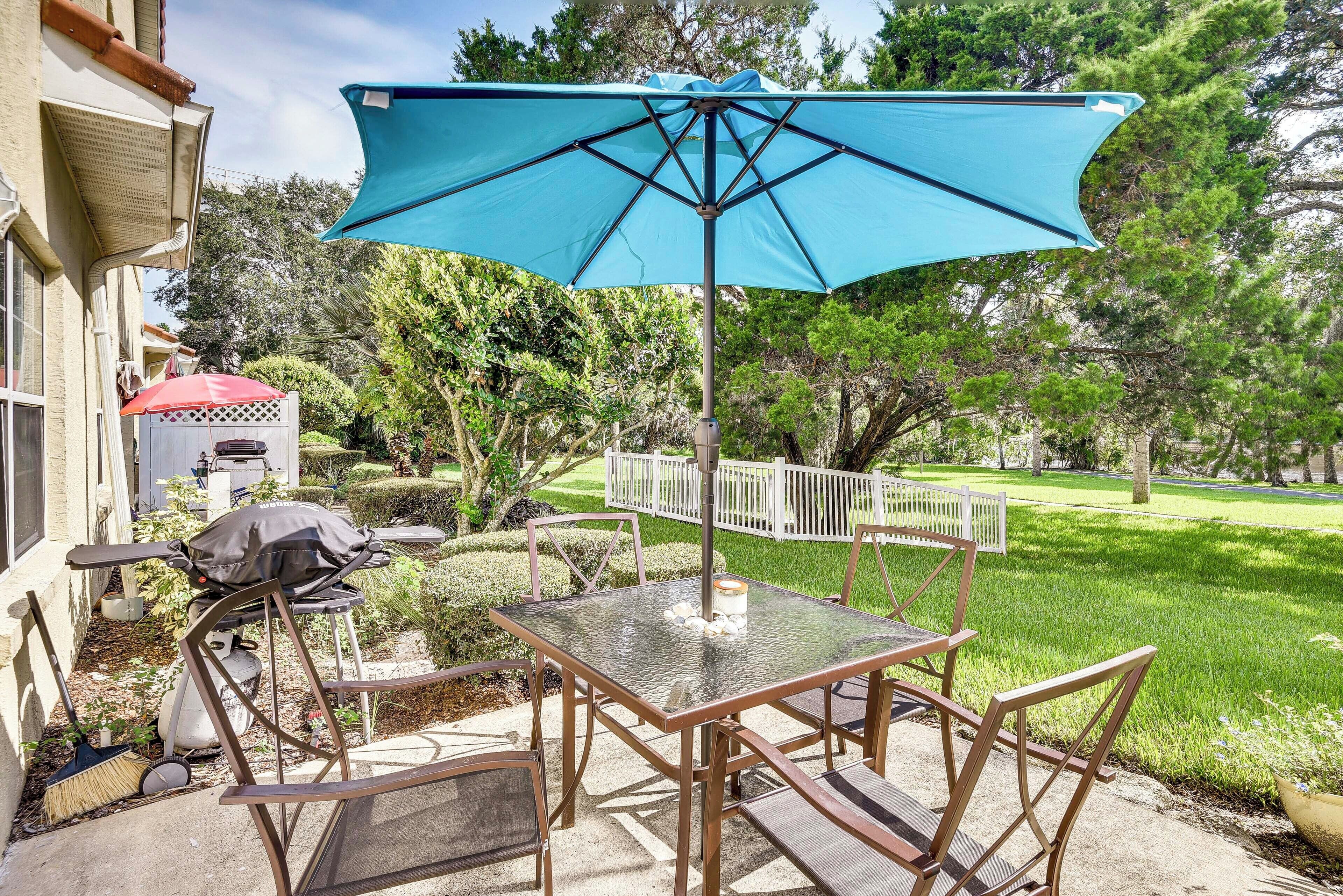 An outdoor patio with a glass table and chairs, a blue umbrella, and a barbecue grill.