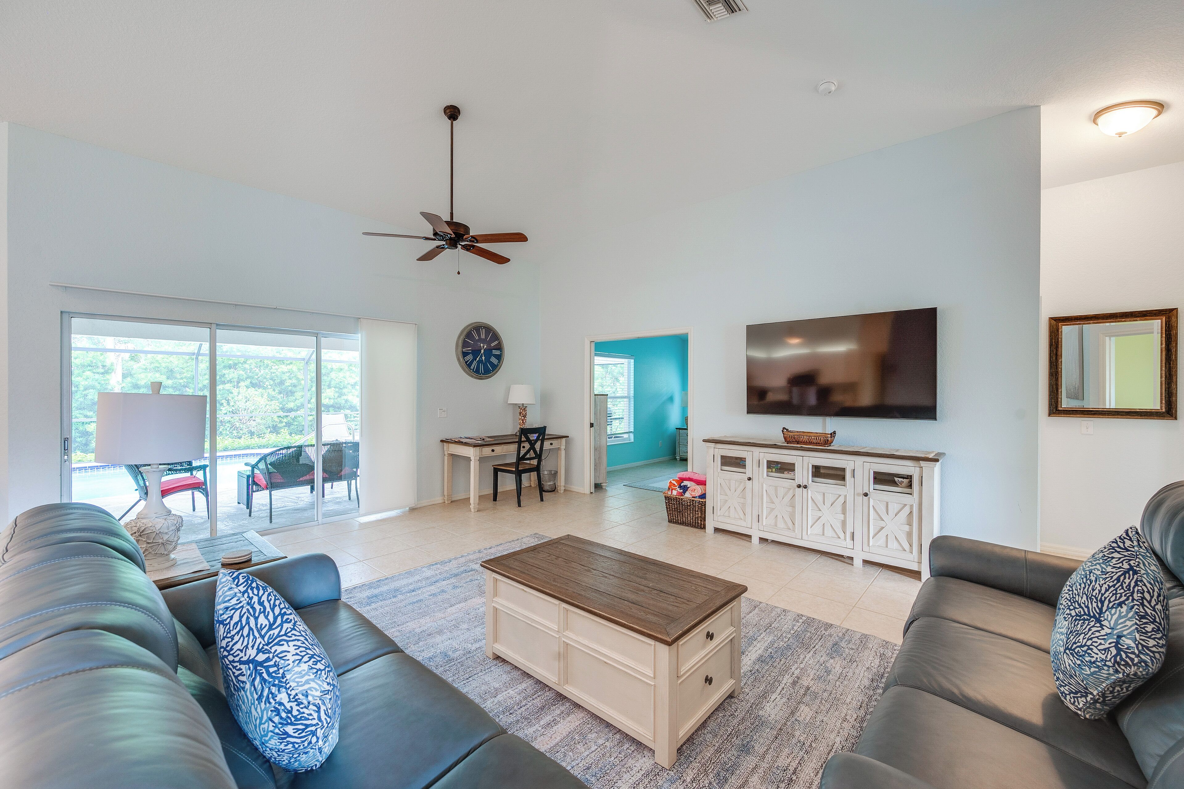 A modern living room with a sofa, coffee table, and TV. There is a dining area with a table and chairs, and a kitchen area with cabinets and a TV.