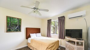 Un dormitorio con una cama, ventilador de techo, aire acondicionado, televisor sobre un mueble bajo y una ventana con cortinas.