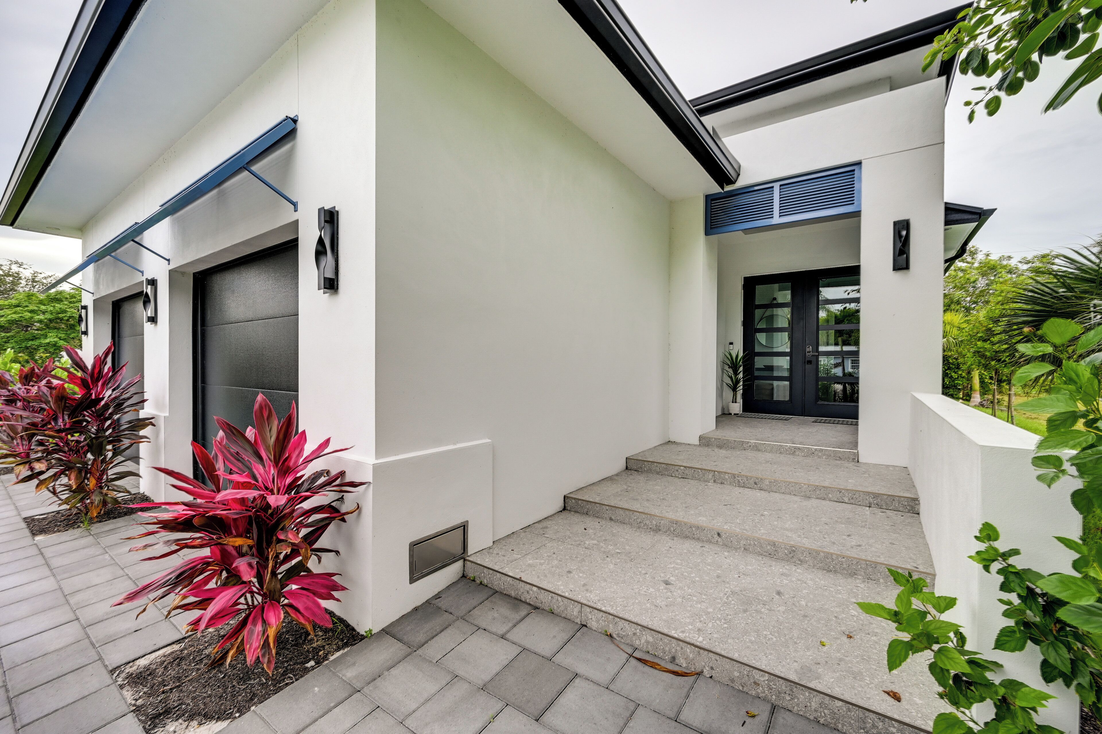 A modern house with a clean design, featuring a large garage door, a set of concrete steps leading to the entrance, and a small garden area with red-leafed plants.