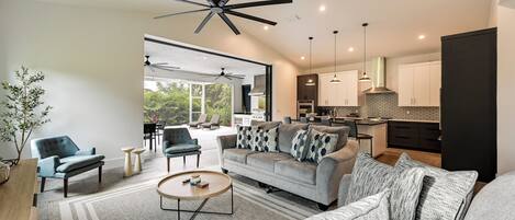 A modern living room with a sofa, armchairs, and a coffee table. There is a kitchen area with cabinets and a dining area visible through an open door.