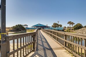 A wooden boardwalk leading to a blue building with a gazebo, surrounded by palm trees and a body of water. - Swim, Soak + Play: Myrtle Beach Resort Condo! (Myrtle Beach)