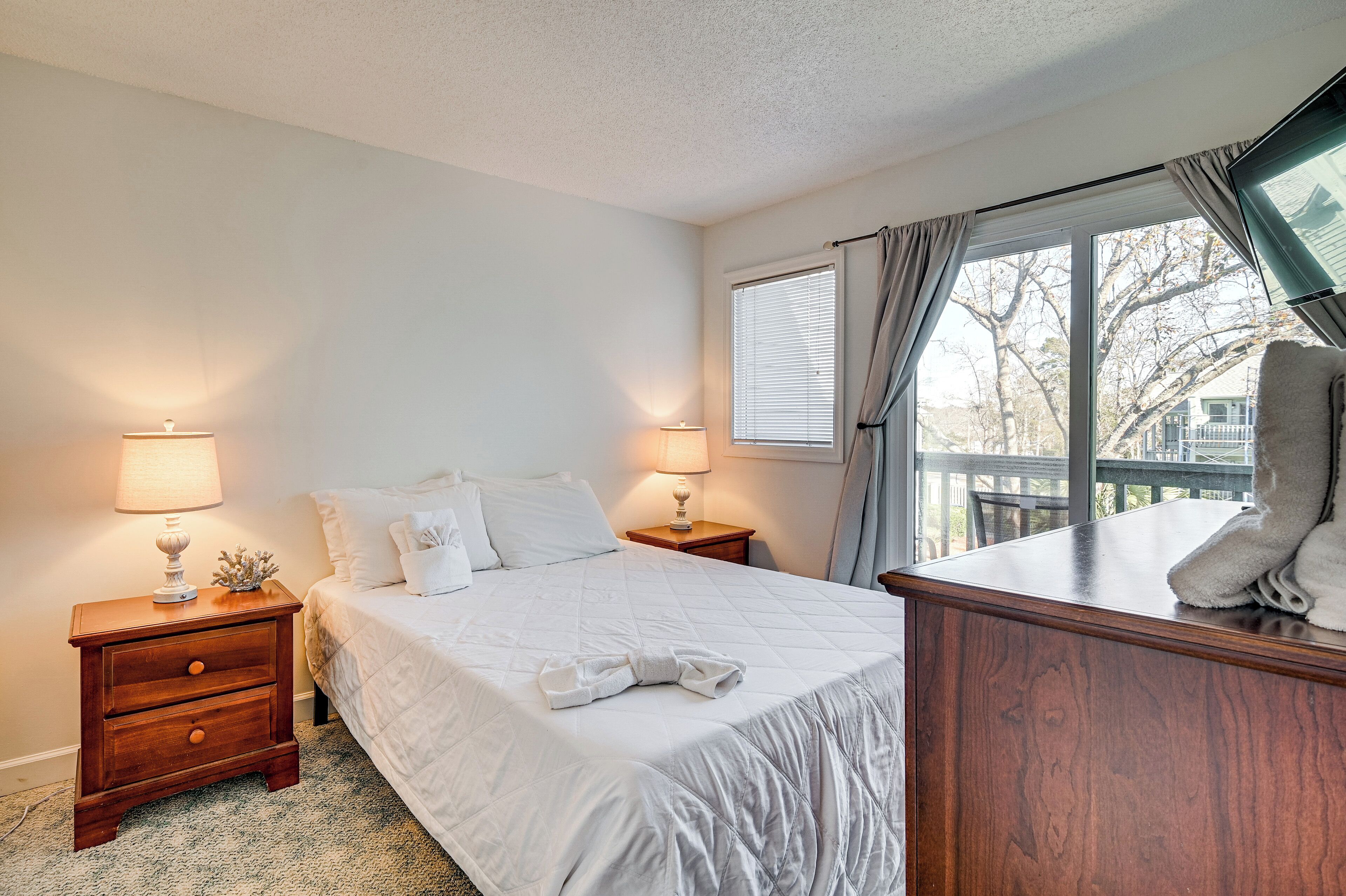 A hotel room with a bed, two bedside tables with lamps, a wooden dresser, and a window with curtains.
