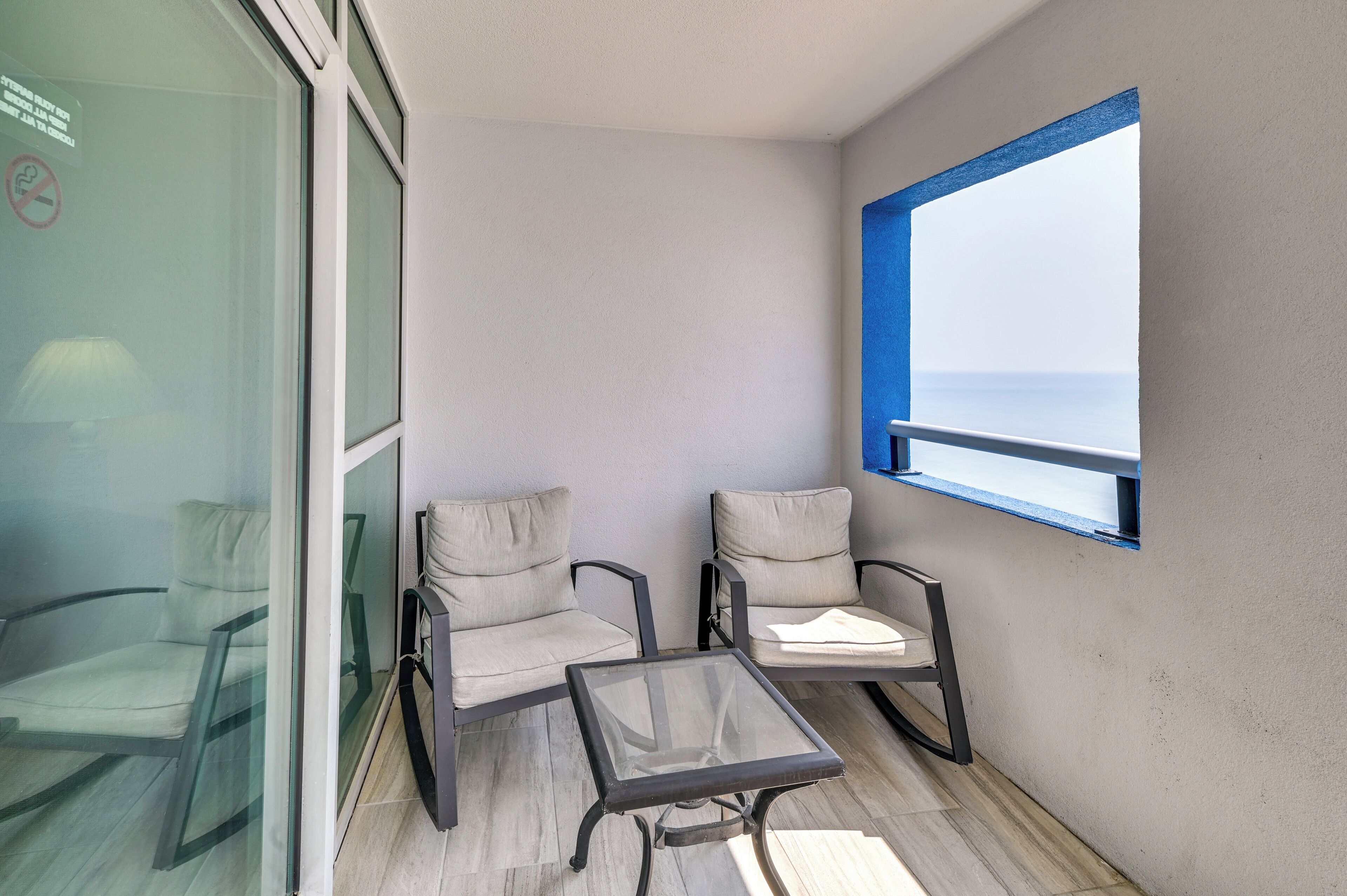 A balcony with two chairs and a small table, a glass door, and a view of the sea through a window.
