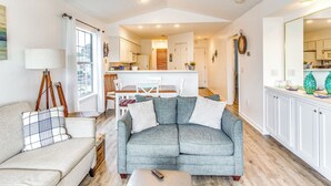 Ein Wohnzimmer mit grauem Sofa, einem weißen Sessel und einem Couchtisch. Im Hintergrund befindet sich eine Küche mit weißen Schränken sowie ein Essbereich mit Tisch und Stühlen.