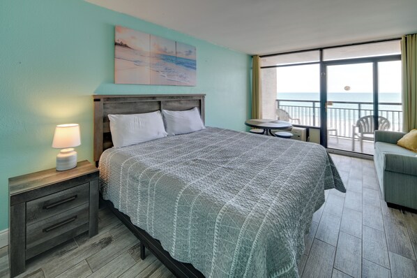Habitación de hotel con cama, mesita de noche, lámpara, un cuadro, balcón con mesa y sillas, y vista al océano.