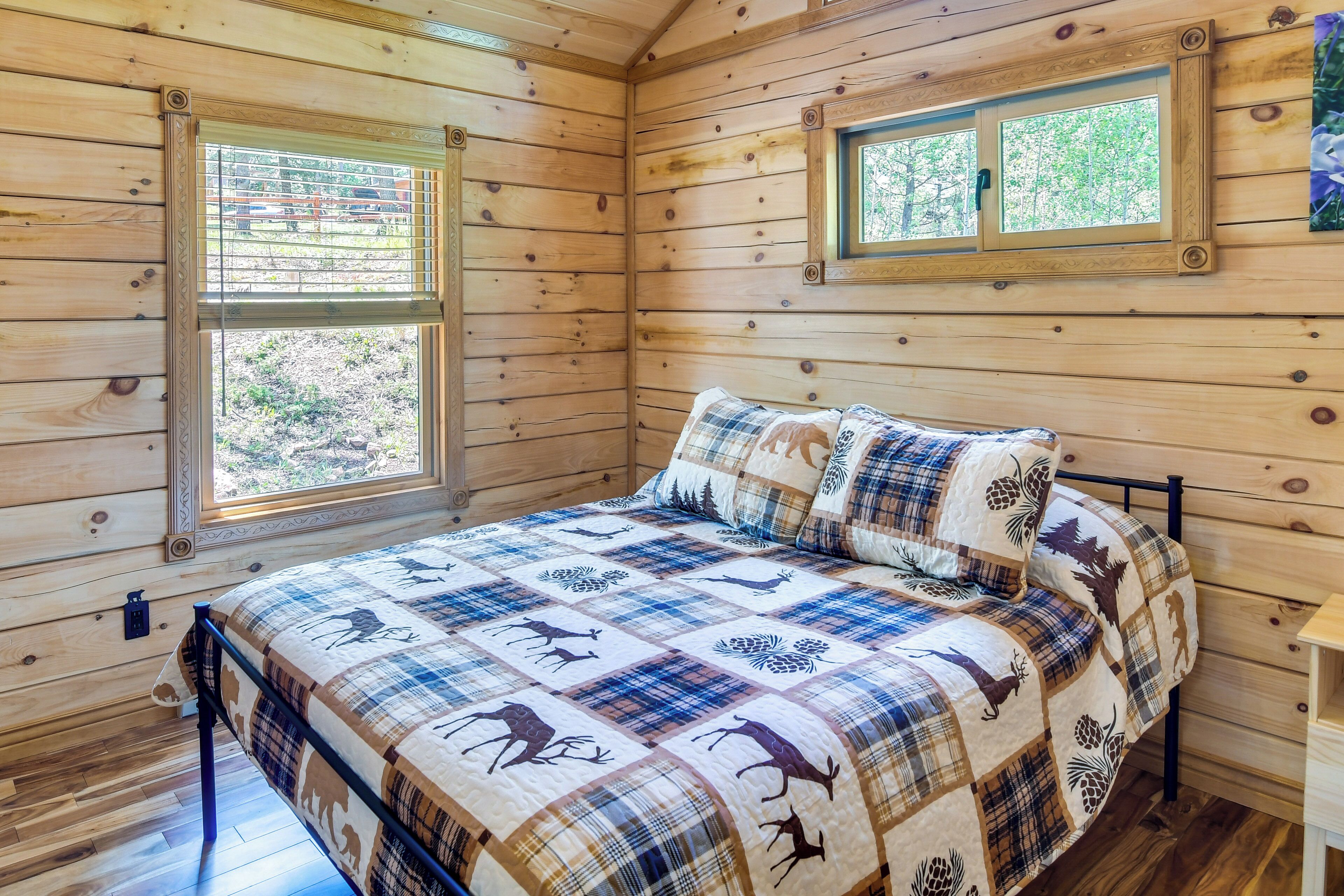 A wooden cabin bedroom with a bed, two windows, and a view of trees.