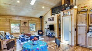 Un interior de cabaña acogedor con piso de madera, una mesa de comedor con mantel floral, chimenea y armarios de madera.