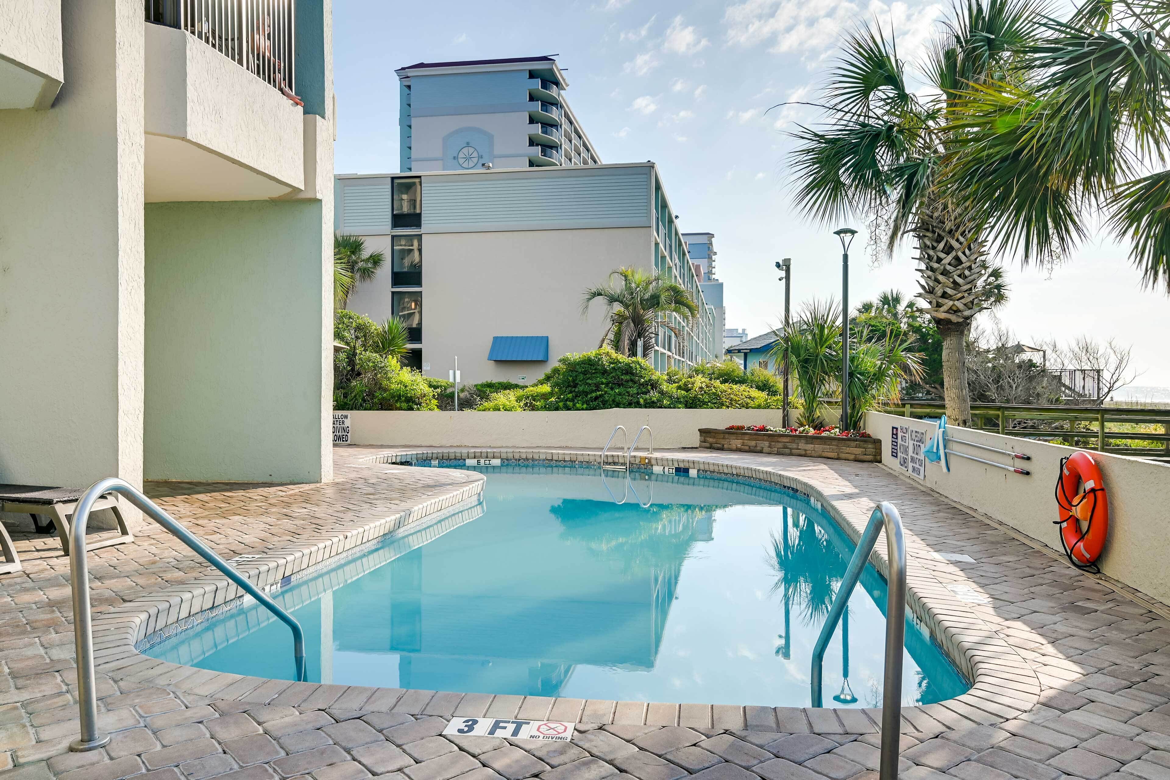 A swimming pool with a 3 FT no diving sign, surrounded by a brick patio, palm trees, and a building with balconies in the background.