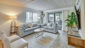 Un salon moderne avec un canapé d’angle gris, un fauteuil blanc, une table basse et un lampadaire. De grandes fenêtres sont équipées de stores, et on y trouve une plante en pot ainsi qu’une porte-fenêtre coulissante donnant sur un balcon.