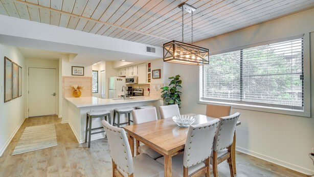 Une cuisine moderne avec des meubles blancs, une table à manger en bois et six chaises, ainsi qu’un luminaire suspendu.