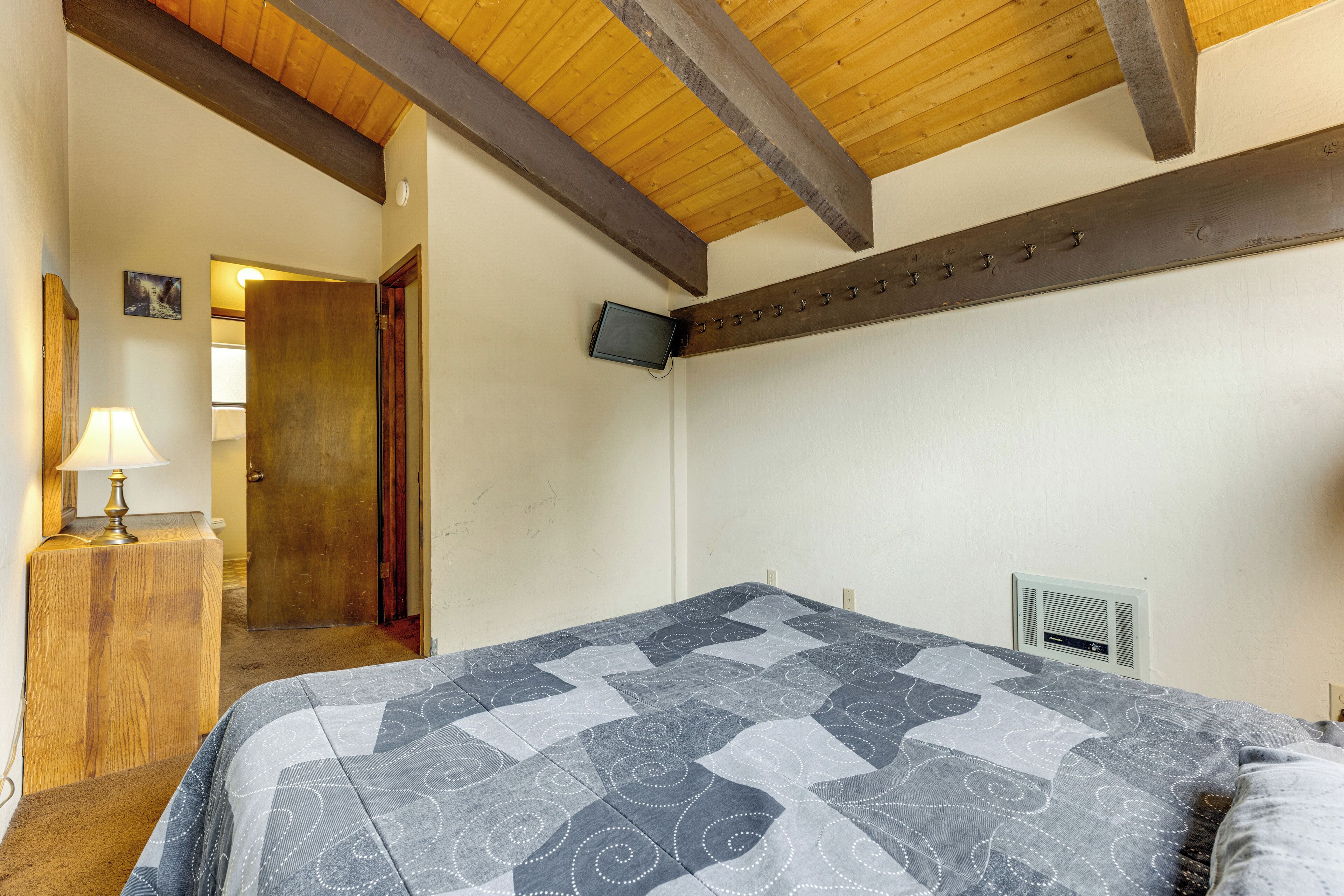 A bedroom with a bed, a wooden dresser, a television mounted on the wall, and a ceiling with exposed beams.