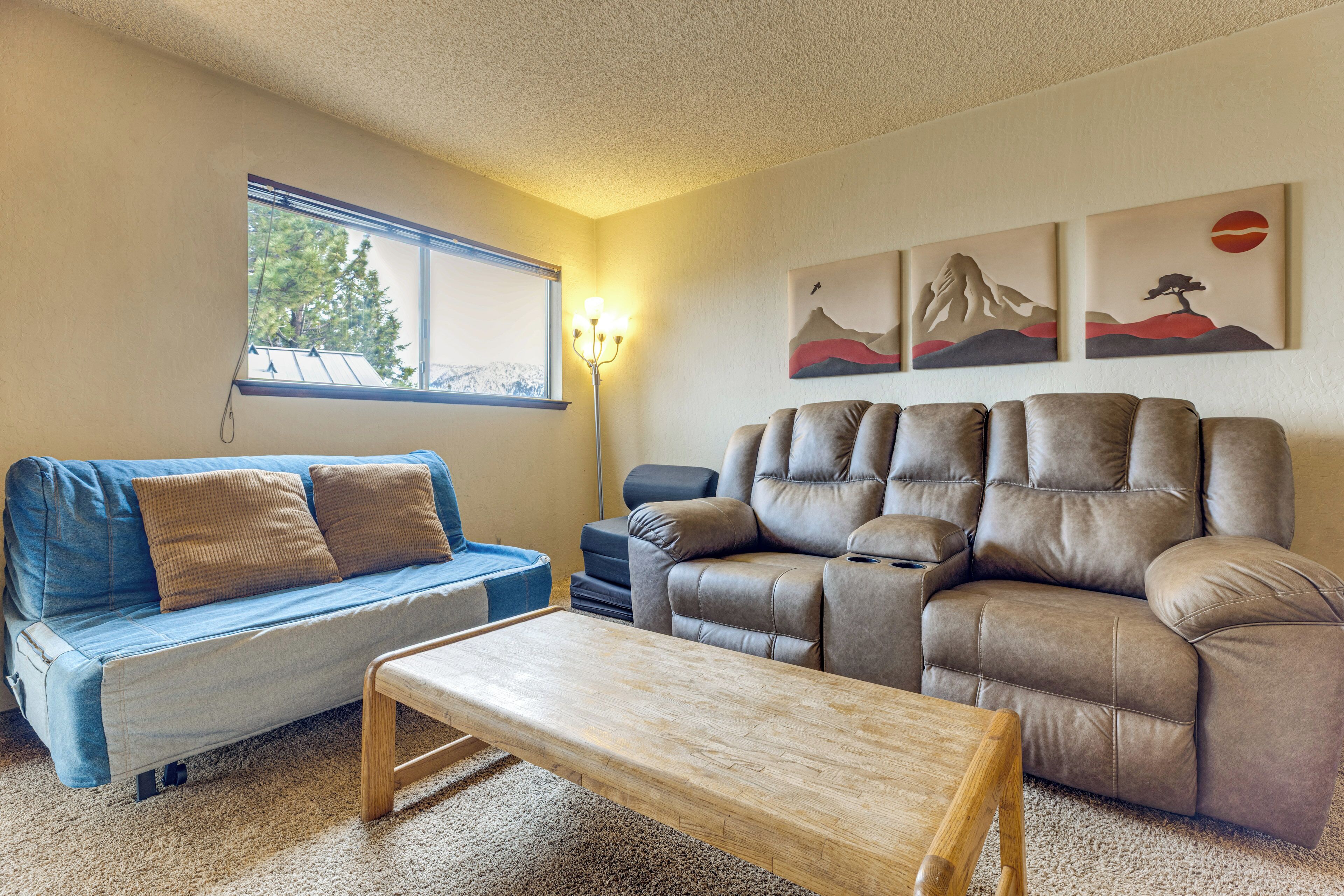 A living room with a blue sofa, a brown leather couch, a wooden coffee table, and wall art depicting mountains and trees.
