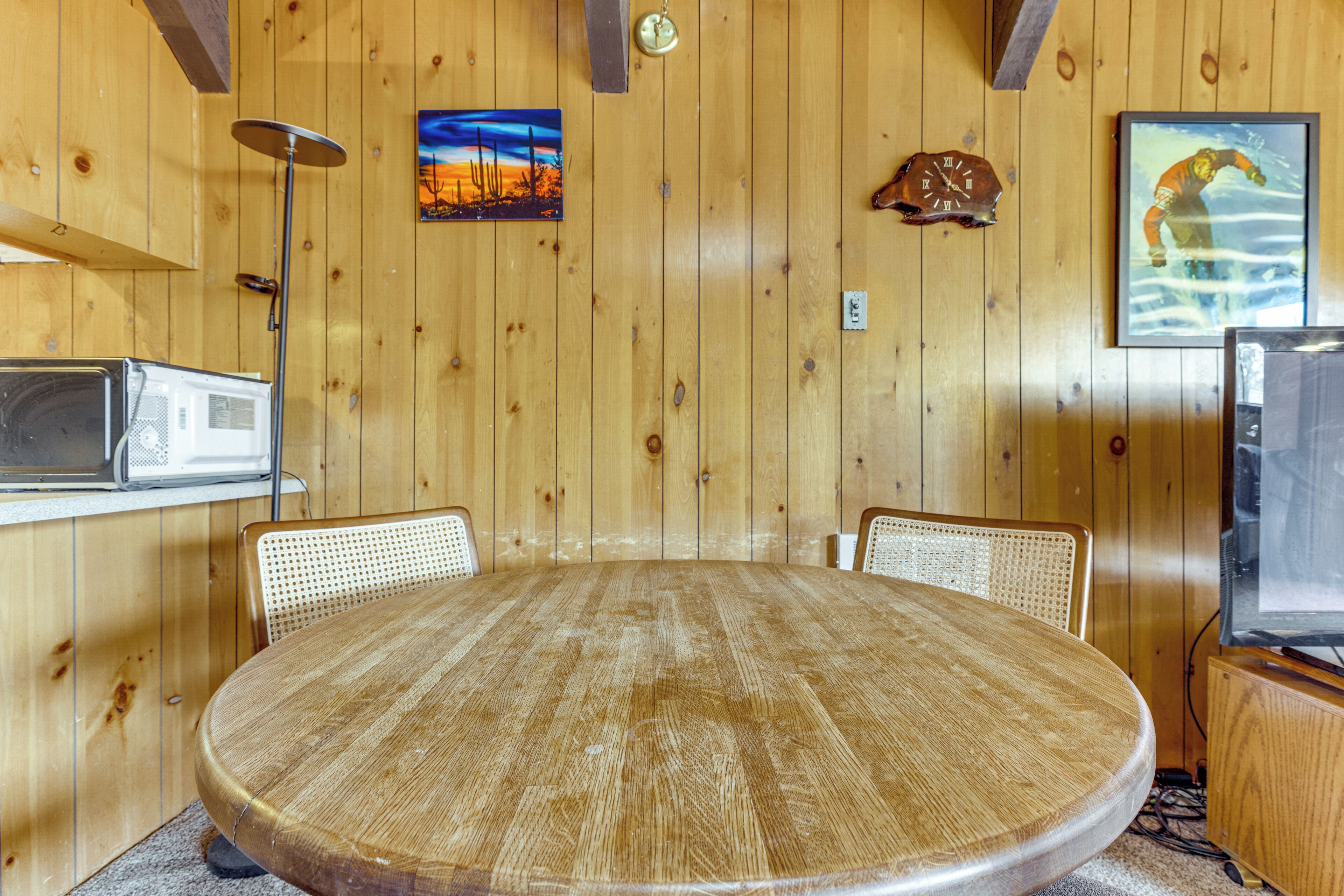 A wooden-paneled room with a round table, chairs, a clock, a framed picture, and a television.