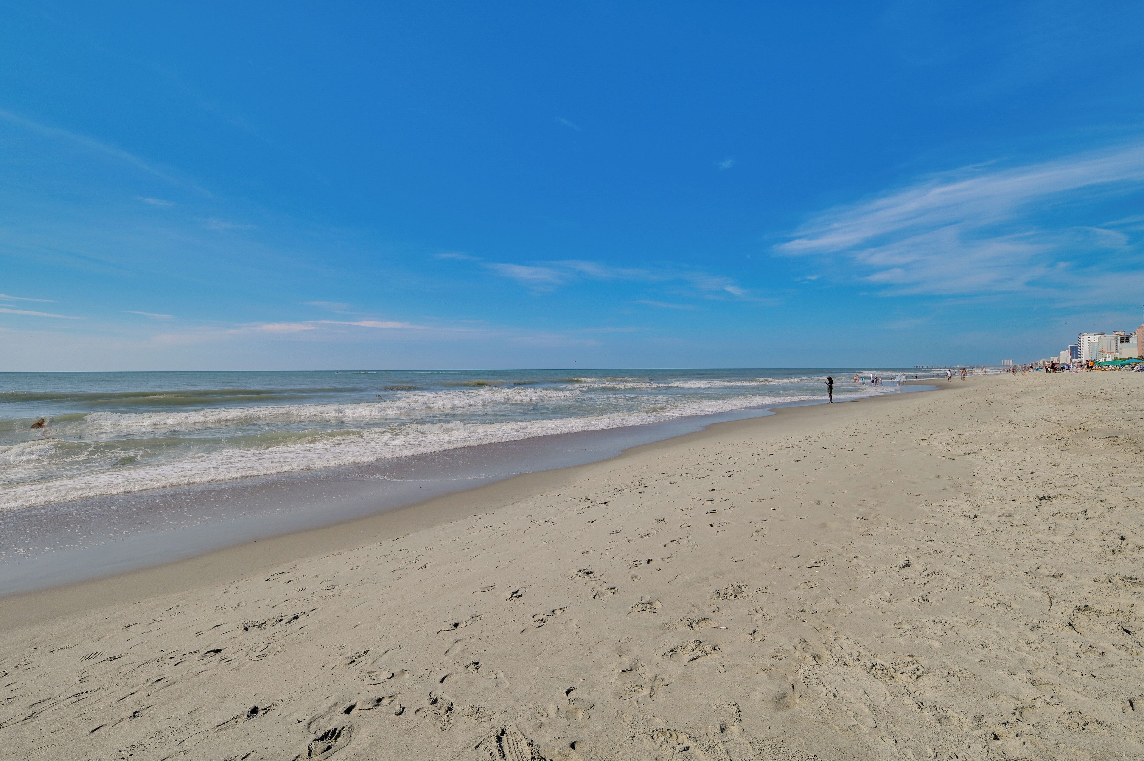 Une vaste plage de sable fin, avec de douces vagues, un ciel dégagé et quelques bâtiments visibles au loin.