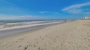 A wide sandy beach with gentle waves, a clear sky, and a few buildings in the background. - Pool Access: Oceanfront Myrtle Beach Getaway! (Myrtle Beach)