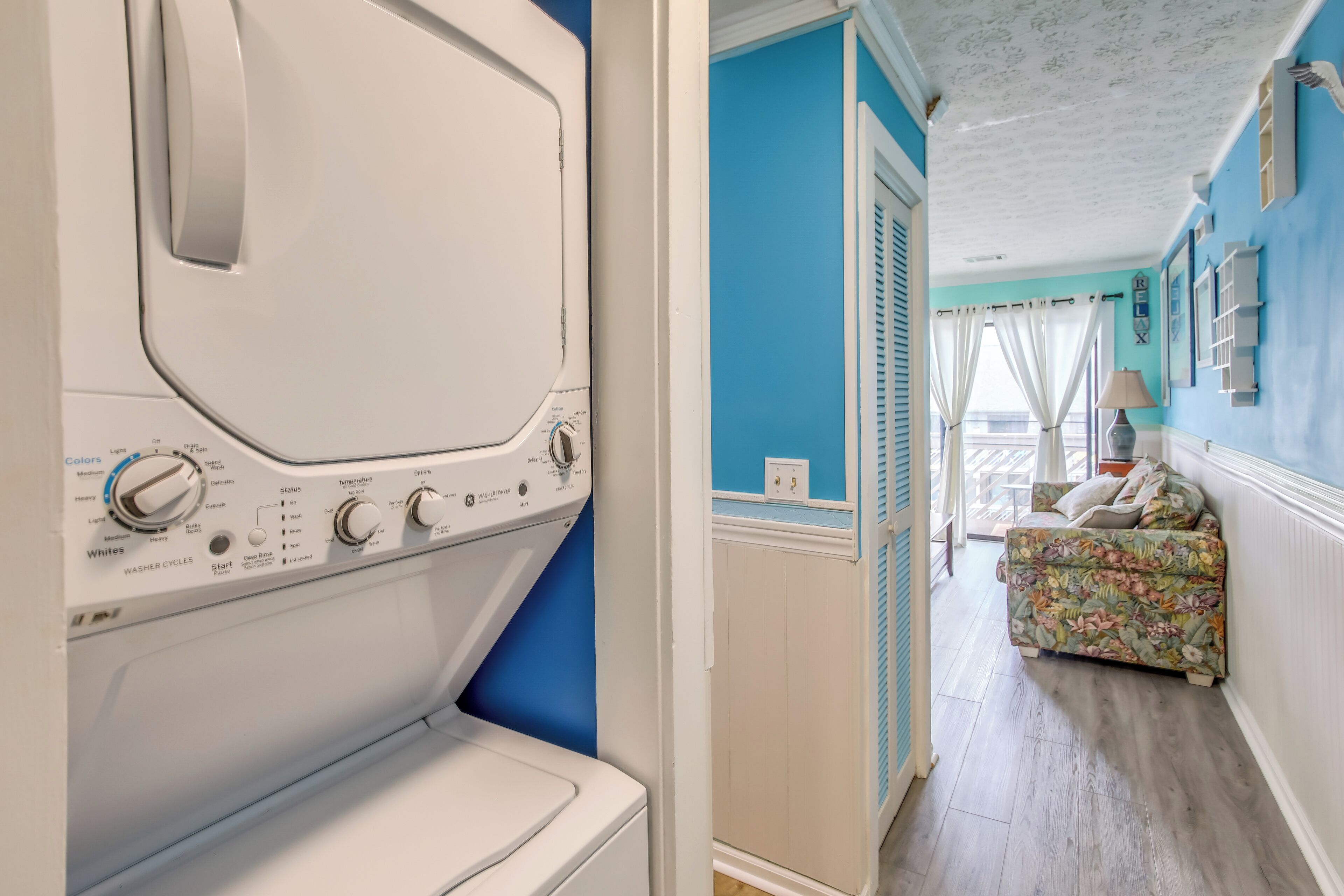 Un área de lavandería con lavadora y secadora de carga frontal, un sofá con estampado floral en una habitación con paredes azules y piso de madera.