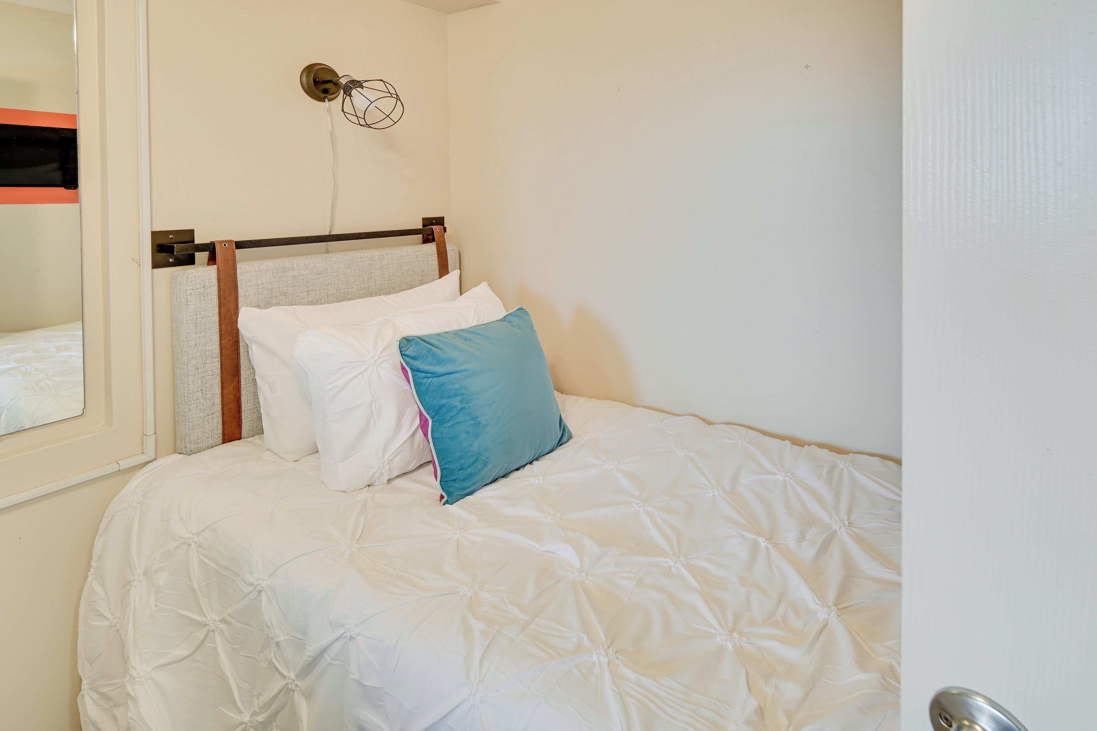 Ein ordentlich bezogenes Bett mit weißer Decke, blauem Kopfkissen, einem Holzkopfteil und einer Wandleuchte.