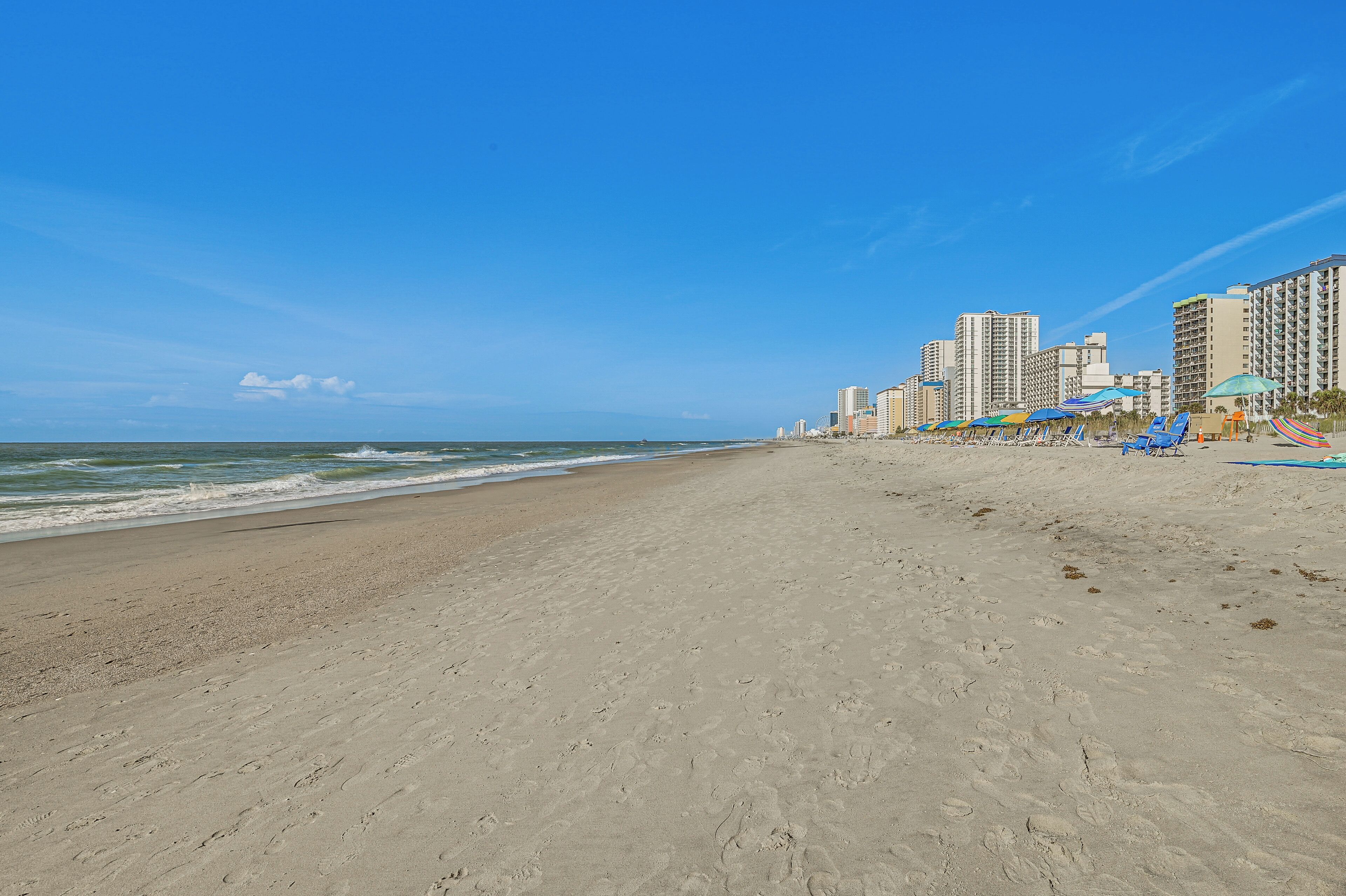 A wide sandy beach with footprints, a clear blue sky, and tall buildings in the background.
