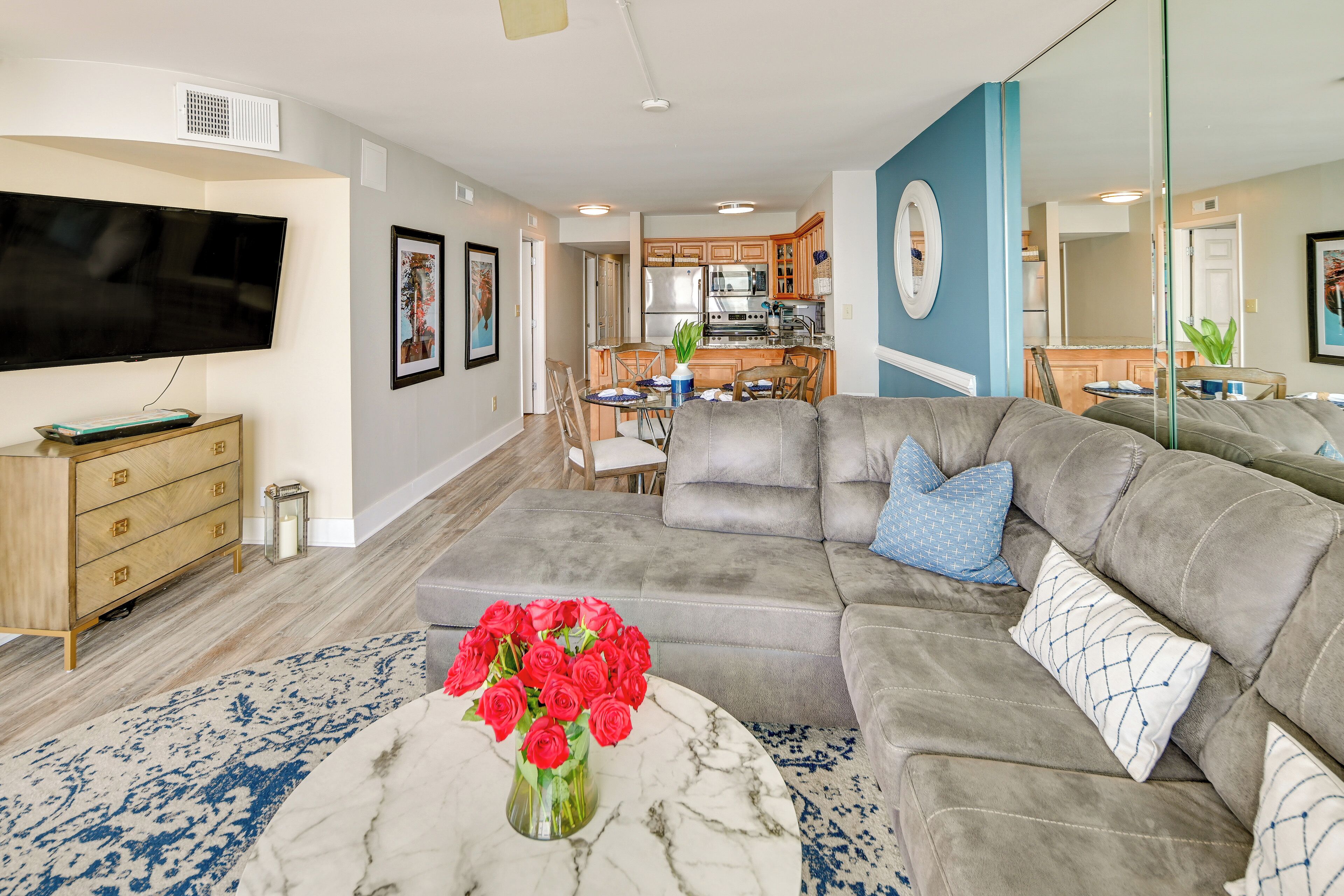 A living room with a grey sectional sofa, a marble coffee table with a vase of red roses, a flat-screen TV, and a kitchen in the background.