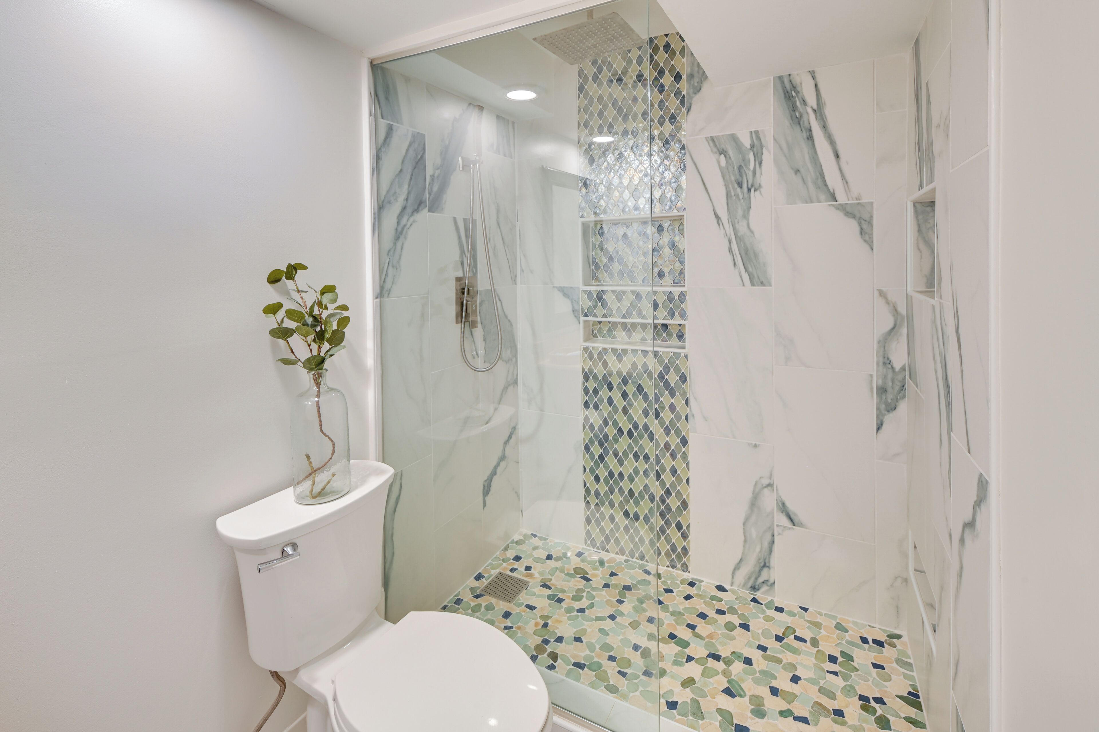 Et moderne badeværelse med en glasafskærmet brusekabine, toilet og mosaikgulv.