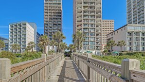 Une promenade en bois mène à de grands immeubles dotés de balcons, le tout étant entouré de verdure.