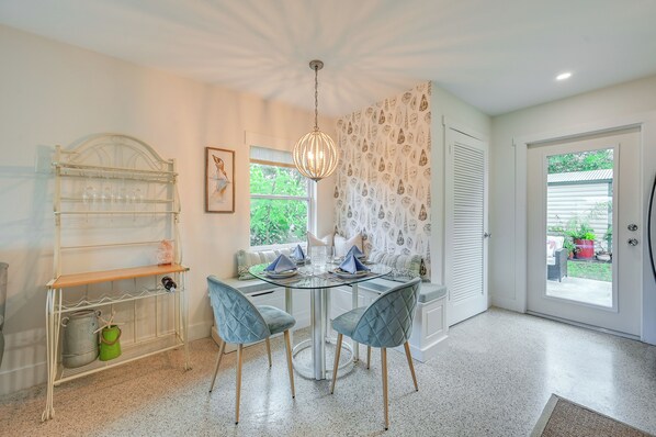 A dining area with a round glass table, four chairs, a hanging light fixture, a white door, and a window with a view of greenery. - Sunny Florida Escape w/ Patio, Grill & Fire Pit! (Naples)